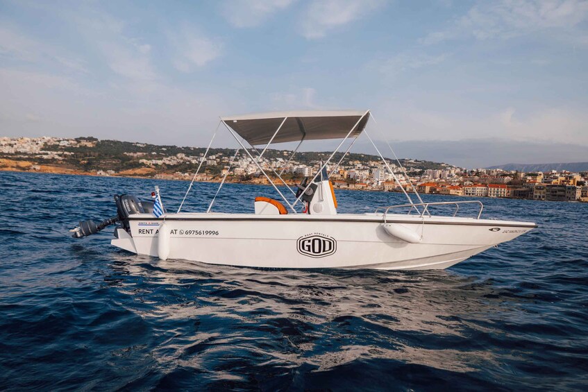 A white motorboat with a canopy, labeled 'GOD,' on calm water with a coastal town in the background. 5