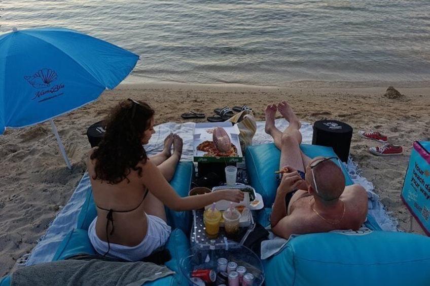 A beach scene with a person lying on a blue lounge chair, another person sitting on a towel, and a third person lying on the sand. 3