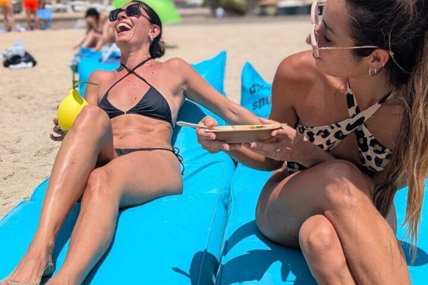 Two people lounging on a blue inflatable lounge chair on a sandy beach. 1