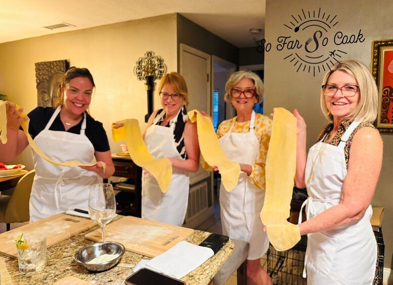Four people in a kitchen holding pasta, with a 'So Far So Cook' sign on the wall. 1