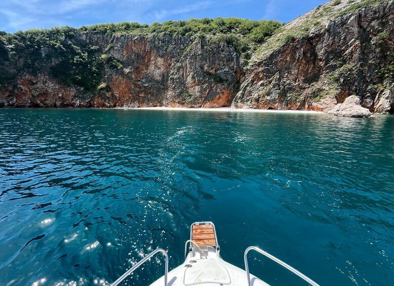 A boat on clear turquoise water with rocky cliffs and a sandy beach in the background. 1