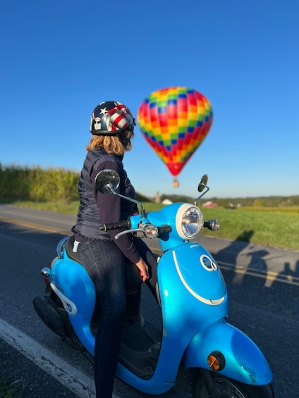A person riding a blue scooter on a road with a colorful hot air balloon in the background. 1