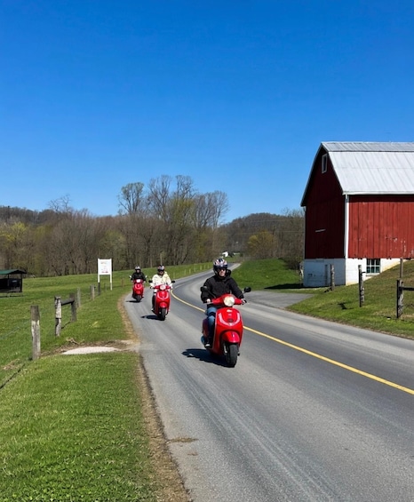 A rural road with three riders on red scooters, a red barn, and a clear blue sky. 3
