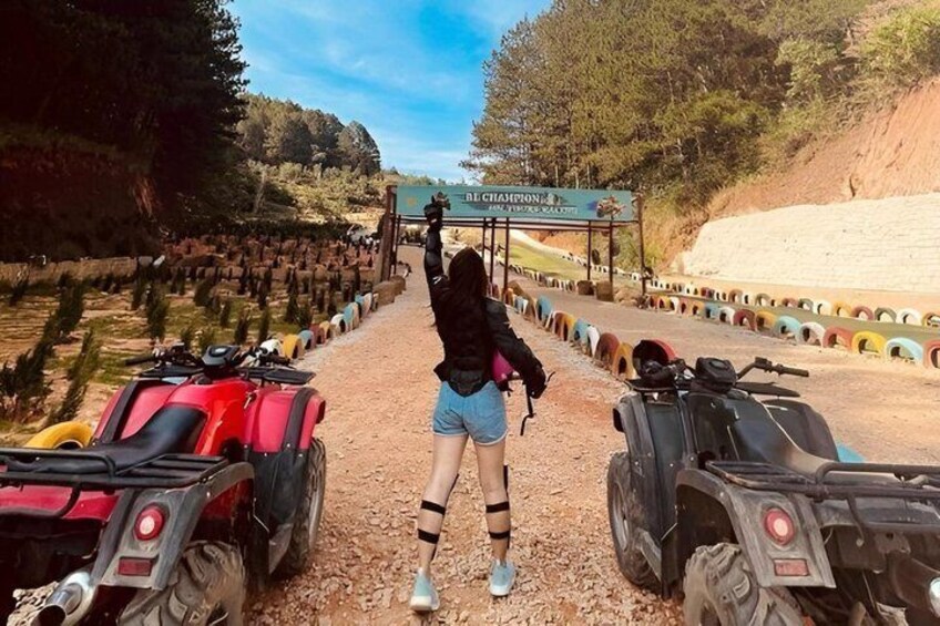A person standing between two quad bikes, a dirt path, and a sign for the 'CHAMPION'S CHALLENGE' in a wooded area. 1