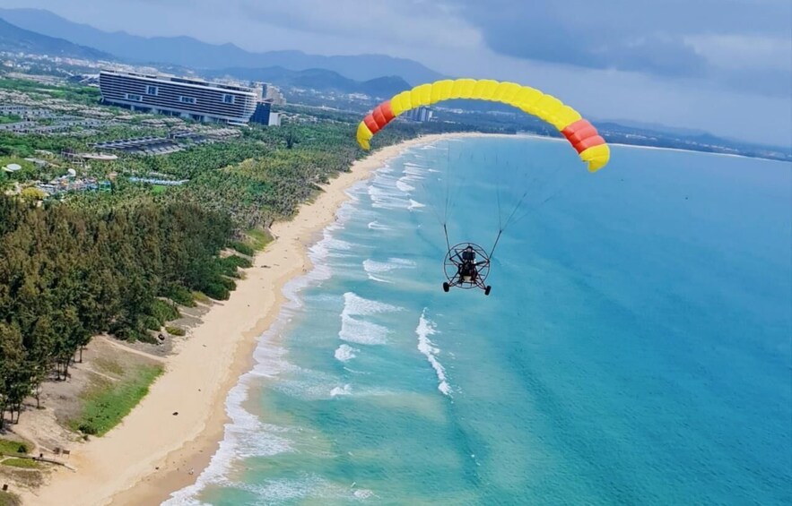 A paraglider flying over a beach with a yellow and red parachute, a cruise ship, and a forested area in the background. 1