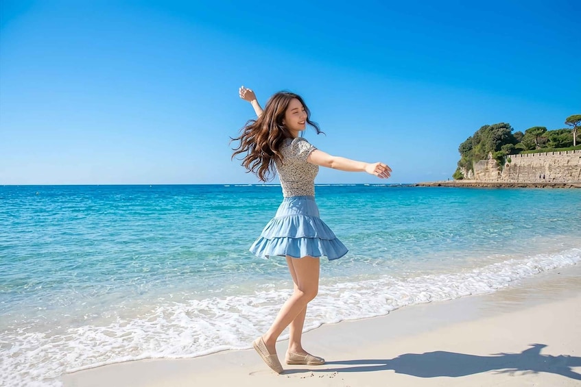 A person dancing on a beach with clear blue water and a stone wall in the background. 1