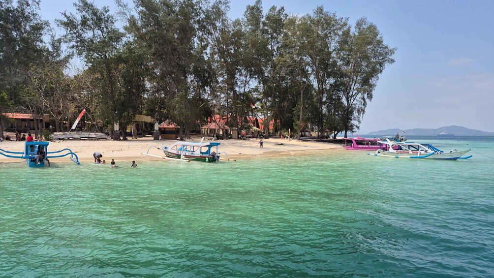 A beach with clear turquoise water, several boats, and a row of trees and buildings in the background. 1