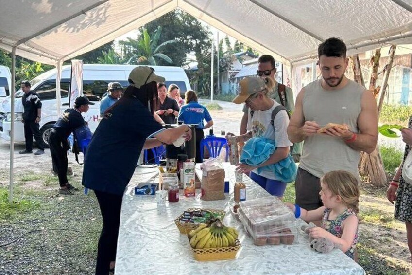 A group of people at an outdoor event with a table displaying food items and a tent overhead. 3