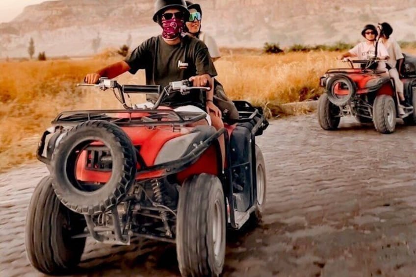 Three people riding ATVs on a cobblestone path in a dry grassland with mountains in the background. 3