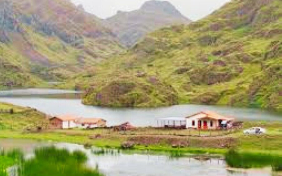 A lake surrounded by hills, a small house with a red roof, and a white car parked by the water. 5