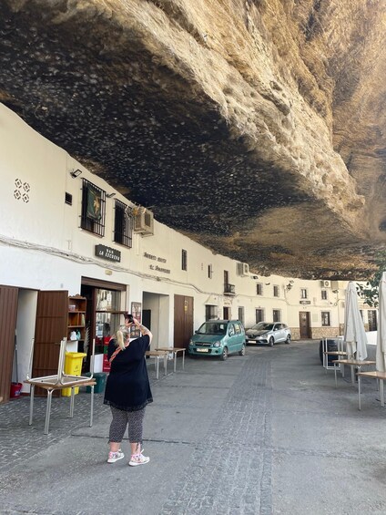 A street with buildings, a parked car, and a person taking a photo. 1