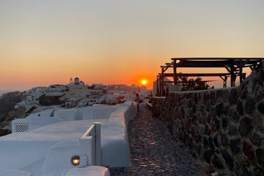 A coastal town at sunset with white buildings, a stone wall, and a wooden pergola. 3