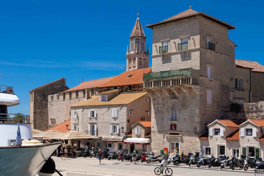 A historic stone building with a tower, a row of shops, and parked scooters. 2