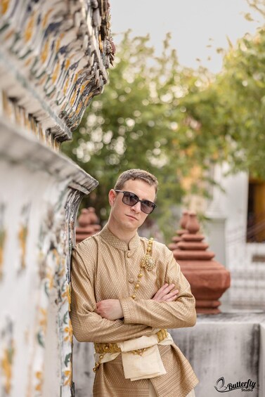 A person in traditional attire standing with arms crossed, leaning against a structure with a patterned design. 2