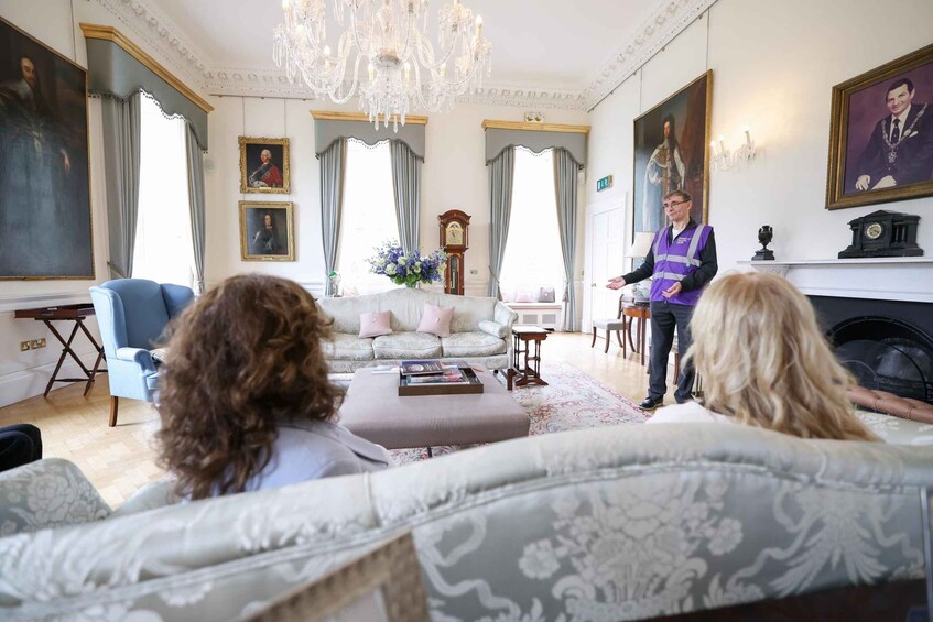 A room with a chandelier, a fireplace, and a person in a purple vest speaking to two seated individuals. 2