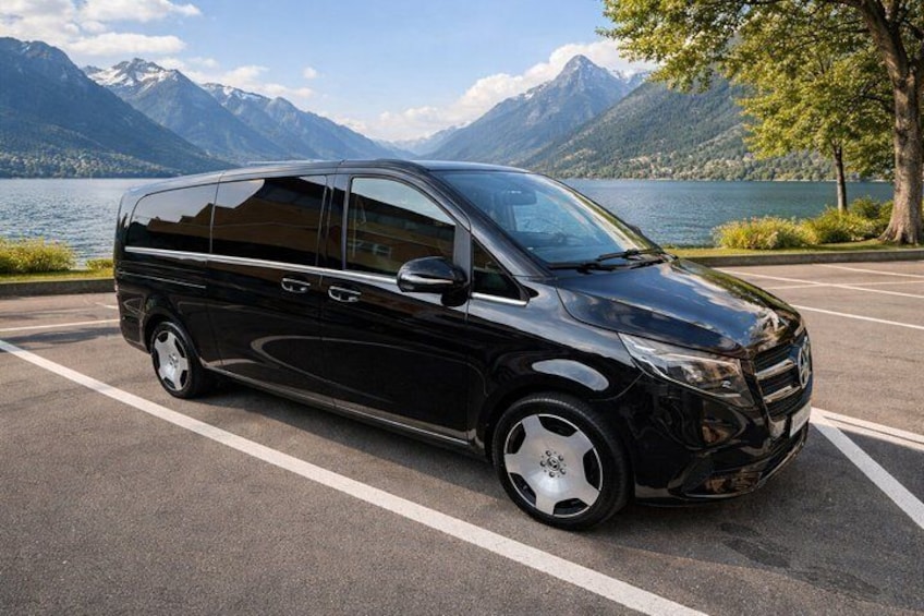 A black minivan parked on a road with a lake and mountains in the background. 2