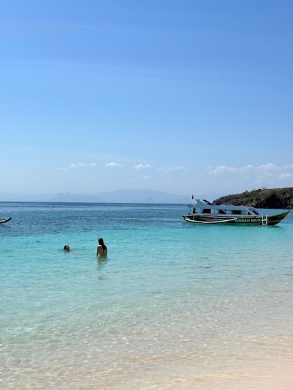 A clear blue sea with two people swimming, a boat named 'ANGIN DARAS' anchored nearby, and distant mountains under a clear sky. 4