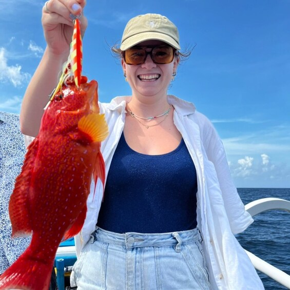 A person holding a red fish, wearing a white shirt, denim shorts, and a cap, standing on a boat with the ocean in the background. 2