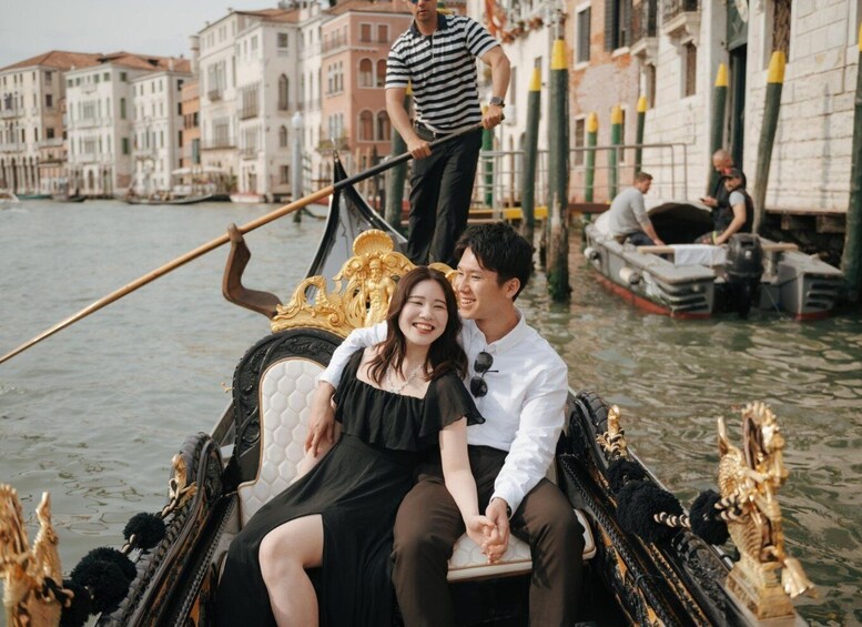 A gondola with ornate decorations on a canal, passengers holding hands, and historic buildings in the background. 1