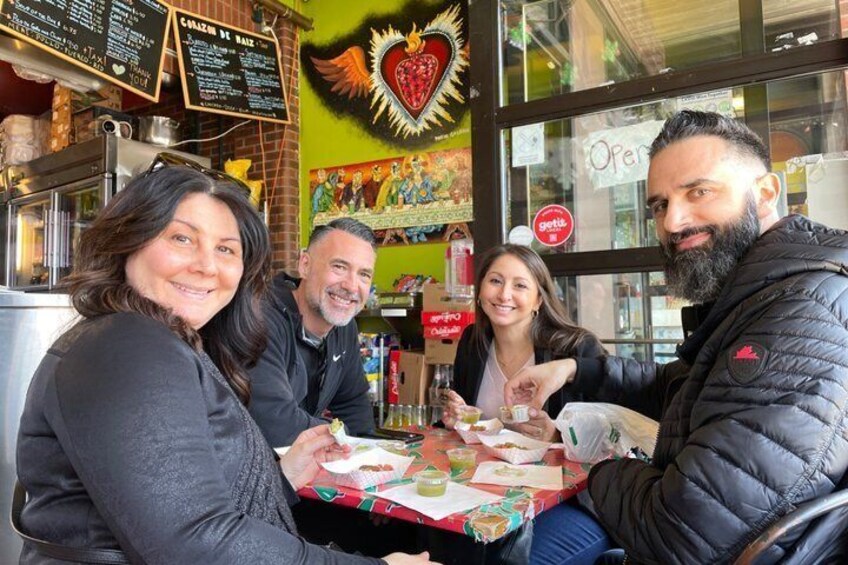 A group of people sitting at a table with food and drinks, in a restaurant with a colorful mural and a menu board. 2