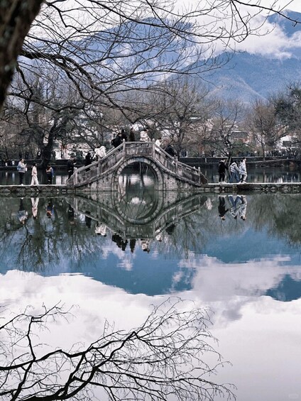 A stone bridge over a calm body of water with reflections, surrounded by leafless trees and mountains in the background. 2