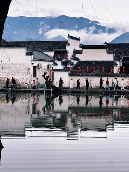 A traditional courtyard with people walking, a calm water body reflecting the scene, and mountains in the background. 5