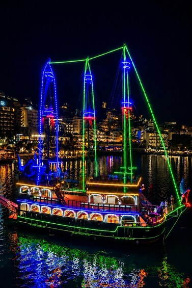 A brightly lit boat with colorful lights, docked at a harbor with buildings in the background. 1