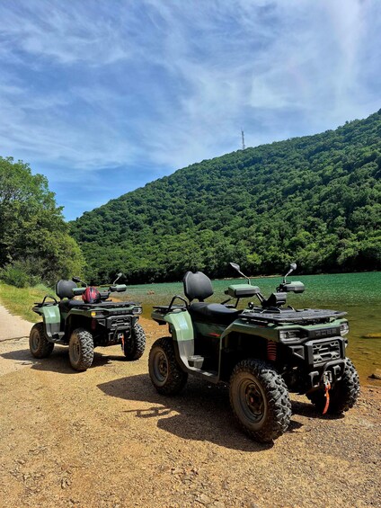 Two ATVs parked on a gravel path with a lake and forested hill in the background. 3