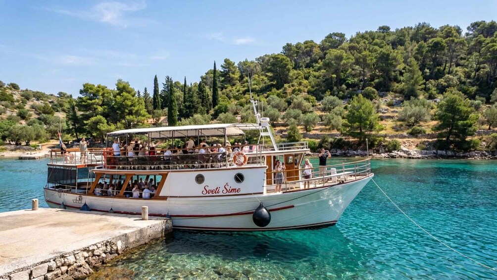 A boat named 'Sveti Sime' docked at a pier with clear turquoise water and a backdrop of dense green trees. 5