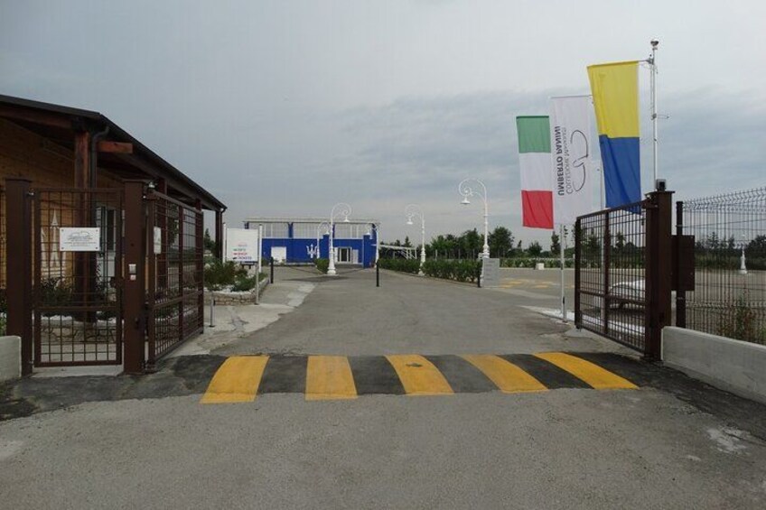 A gated entrance with a blue building in the background, a yellow and black striped crossing, and flags on poles. 2