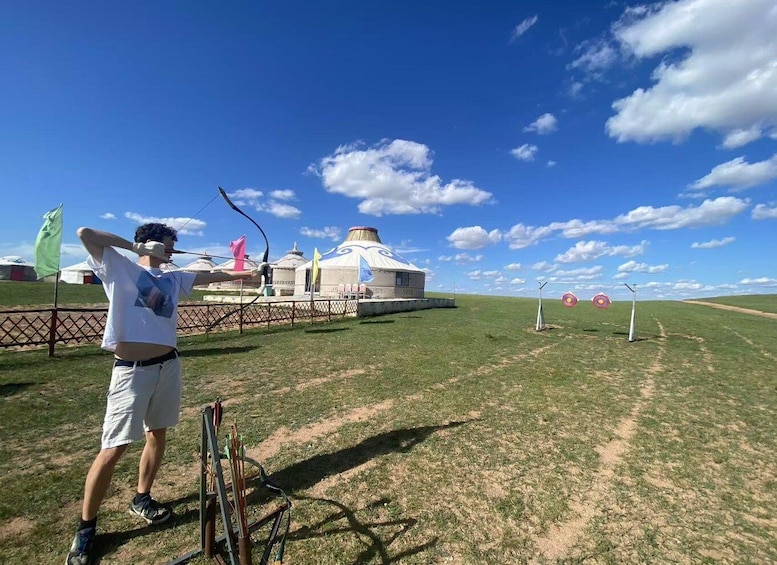 A person aiming an arrow at a target in an open field with yurts in the background. 2