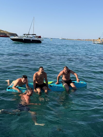 Four people on surfboards in the water with boats and a rocky shoreline in the background. 2