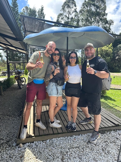 Four people standing under a large umbrella on a wooden deck with gravel ground, trees, and a building in the background. 3