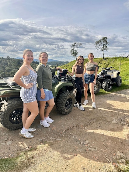 Four people standing on a dirt path with ATVs in the background, surrounded by green hills and a cloudy sky. 5