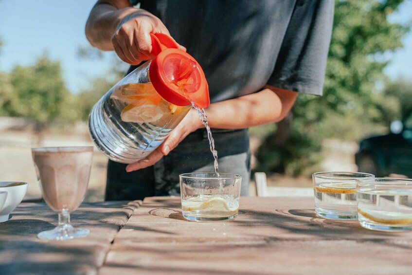 A person pouring a clear liquid from a red-lidded pitcher into a glass on a wooden table. 2