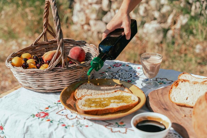 A picnic setup with a basket of fruits, a bottle of honey, slices of bread, a cup of coffee, and a glass of milk on a tablecloth. 3