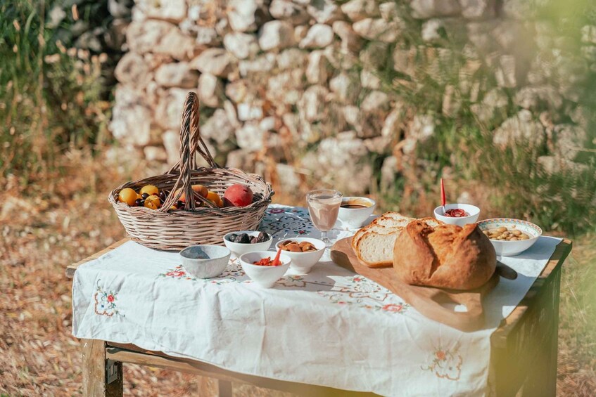 A picnic setup with a basket of fruits, bowls of food, and a loaf of bread on a table outdoors. 1