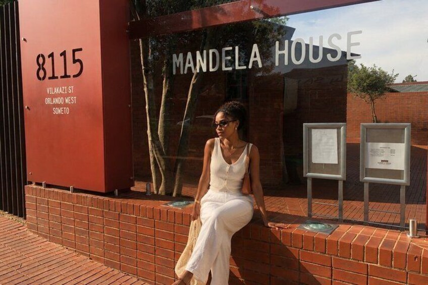 A person sitting on a brick ledge in front of a building labeled 'Mandela House' with a number '8115' and address 'Vilakazi St, Orlando West, Somfeto' visible. 3