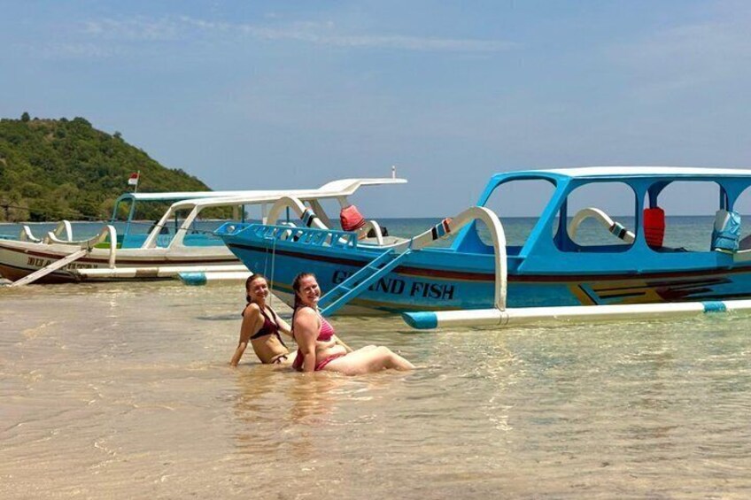 Two people sitting on a beach with boats in the background. 5