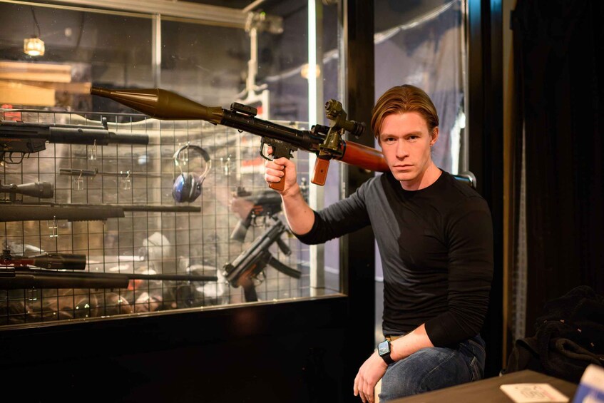 A person holding a rifle with a scope, sitting in front of a display of various firearms. 3
