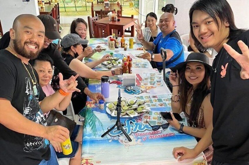 A group of people seated around a table with a beach-themed tablecloth, a tripod, and various items on the table. 1