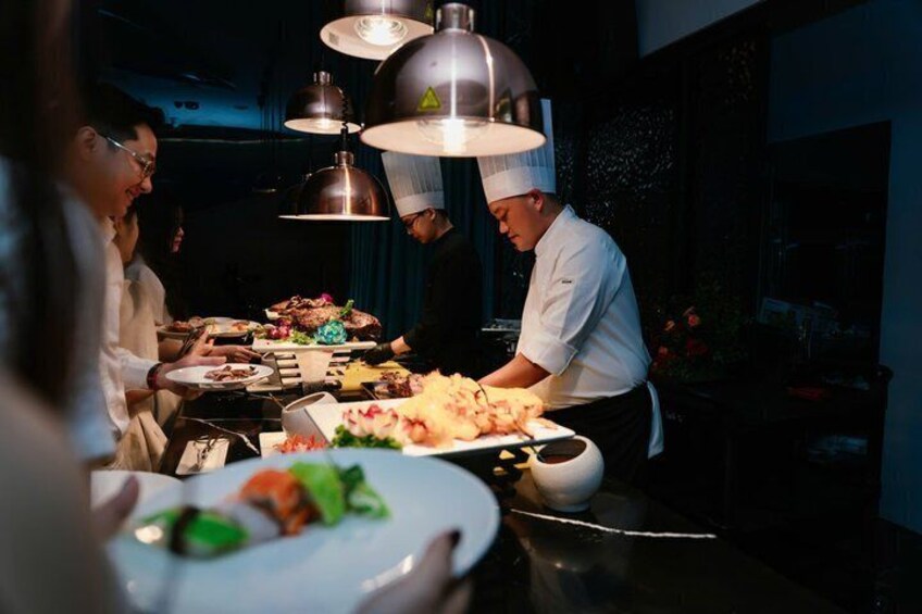 A chef preparing food in a professional kitchen with hanging lights and a dining table in the foreground. 2