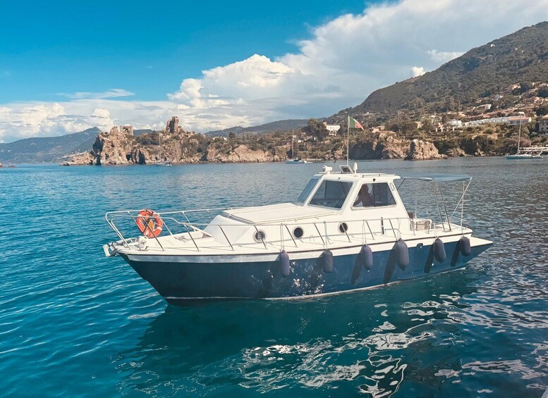 A boat on calm water with a rocky coastline and buildings in the background. 1