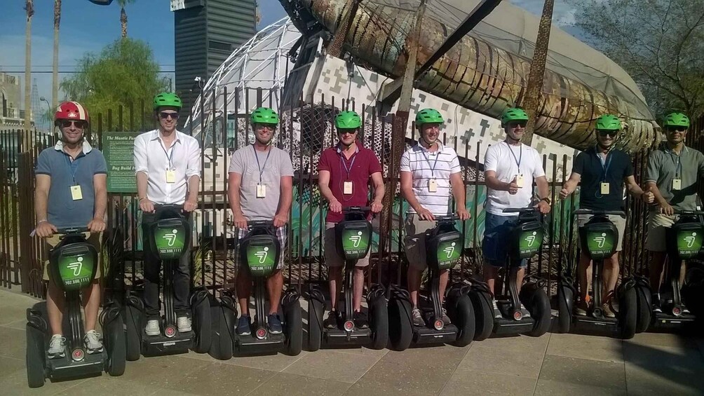 A group of people on Segway devices, wearing helmets, standing in a line outdoors with a large structure in the background. 3