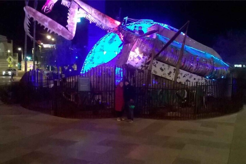 A large illuminated sculpture resembling a fish with a blue and purple color scheme, surrounded by a fence. 4