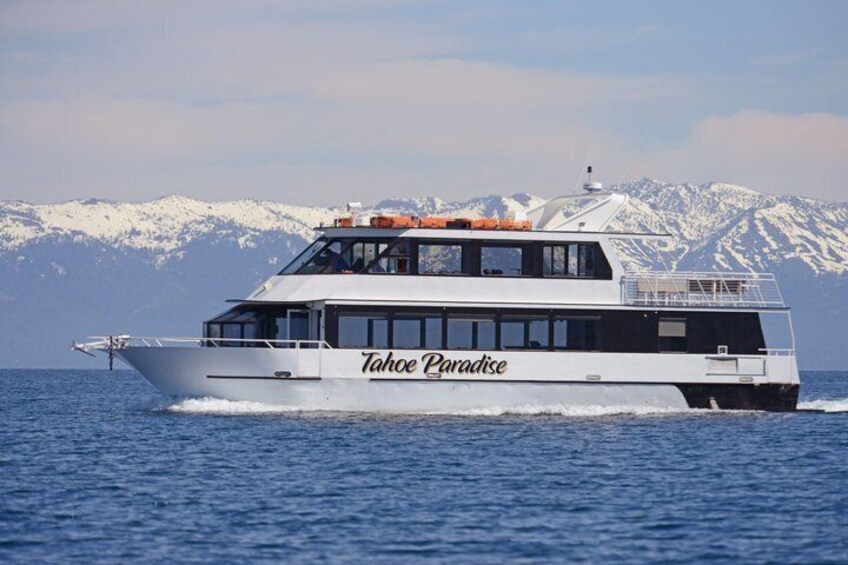 A white boat named Tahoe Paradise on the water with snow-capped mountains in the background. 1