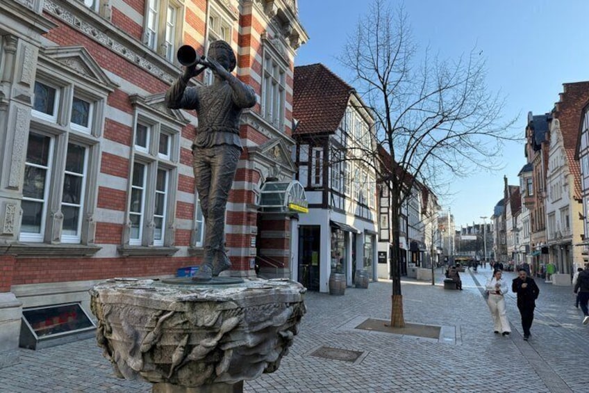 A cobblestone street with a statue of a person playing a trumpet on a pedestal, historic buildings with red brick facades, and pedestrians walking by. 1