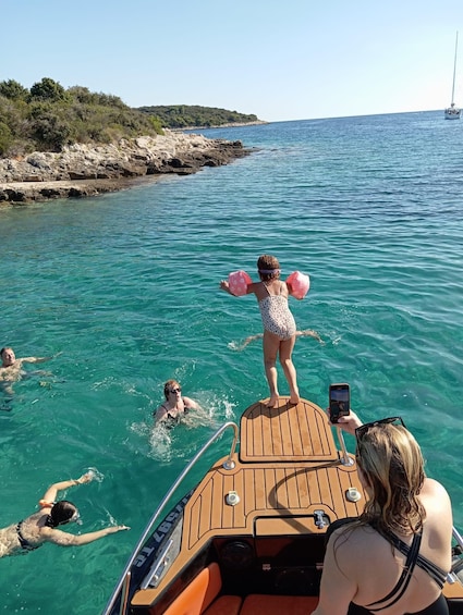 A boat with people swimming and standing in the water, a rocky coastline, and a clear blue sea. 3