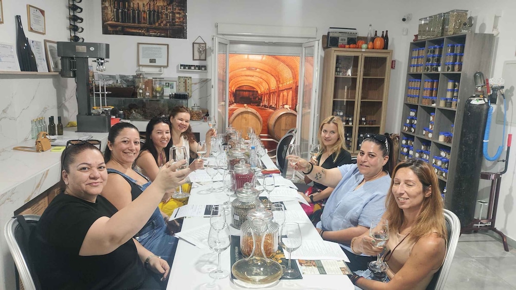 A group of people seated around a table with wine glasses, in a room with wine barrels and shelves of jars. 3