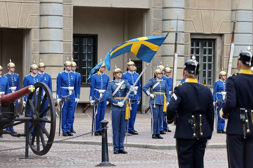 A group of uniformed guards in blue and yellow attire standing in formation with a flag, in front of a building with classical architecture. 3